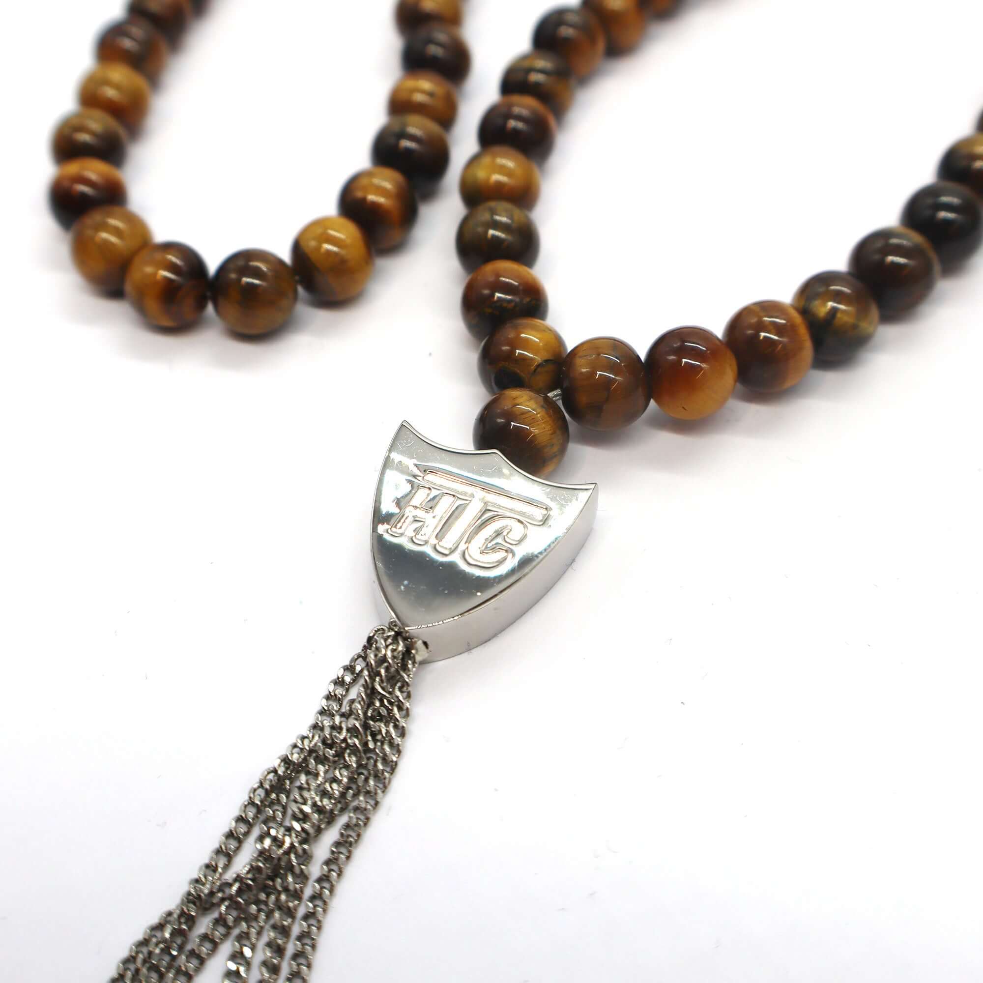 htc los angeles MYSTERY SHIELD NECKLACE TIGER EYE Tiger Eye necklace featuring a blend of textile, glass, and metal materials, with a metal HTC logo detail. Hollywood Trading Company