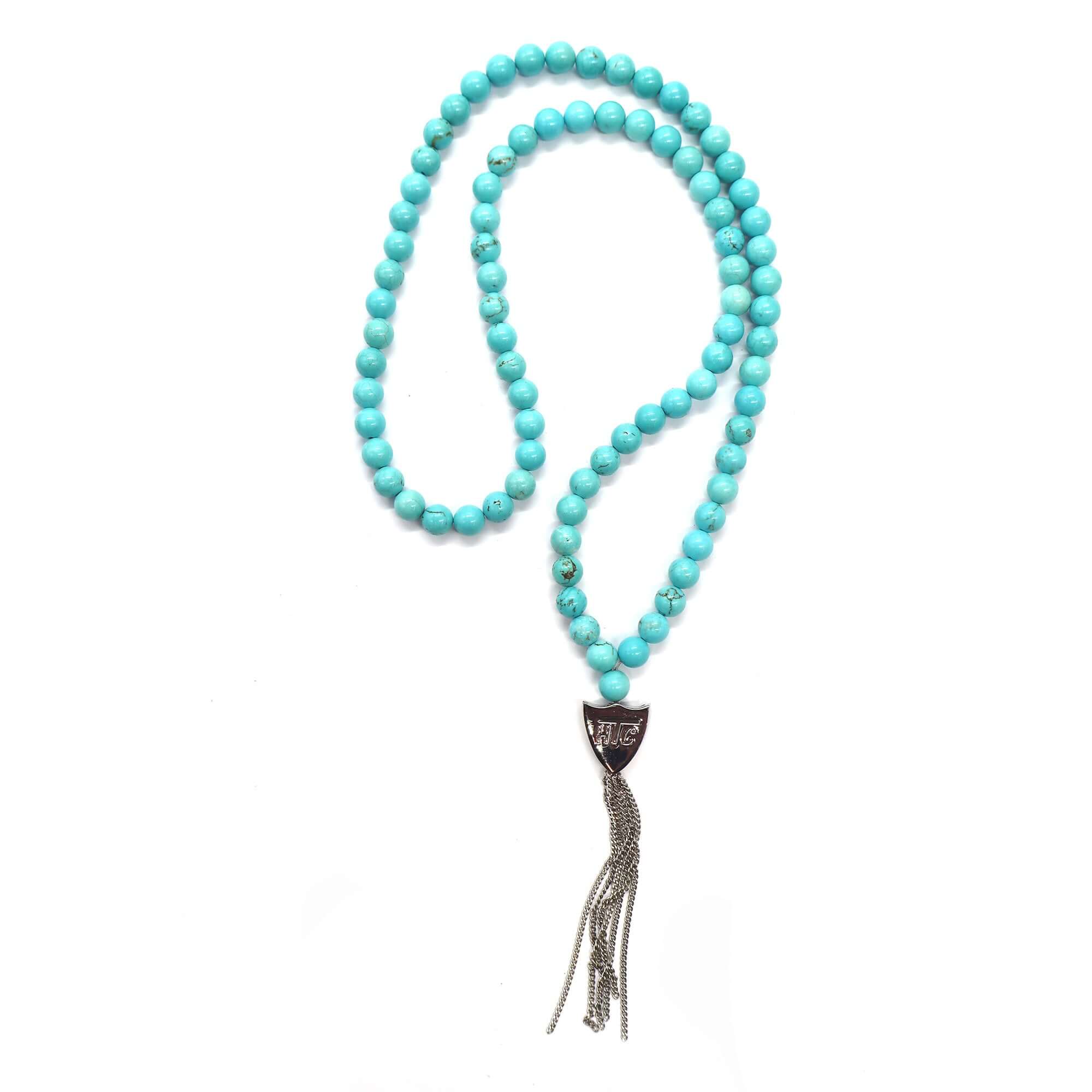 htc los angeles MYSTERY SHIELD NECKLACE TURQUOISE Turquoise necklace featuring a blend of textile, glass, and metal materials, with a metal HTC logo detail. Hollywood Trading Company