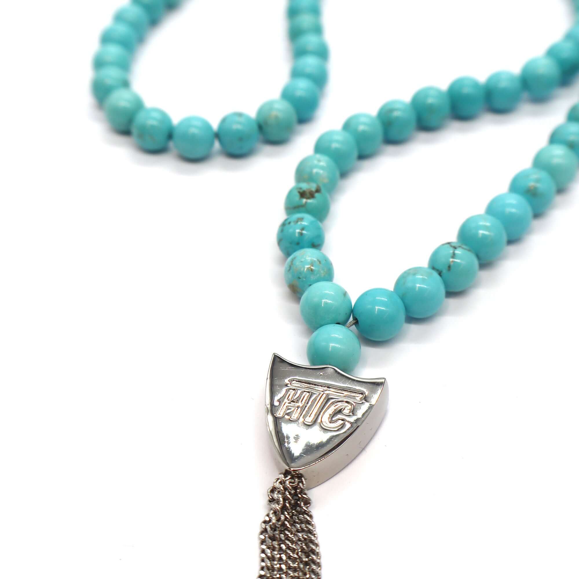 htc los angeles MYSTERY SHIELD NECKLACE TURQUOISE Turquoise necklace featuring a blend of textile, glass, and metal materials, with a metal HTC logo detail. Hollywood Trading Company