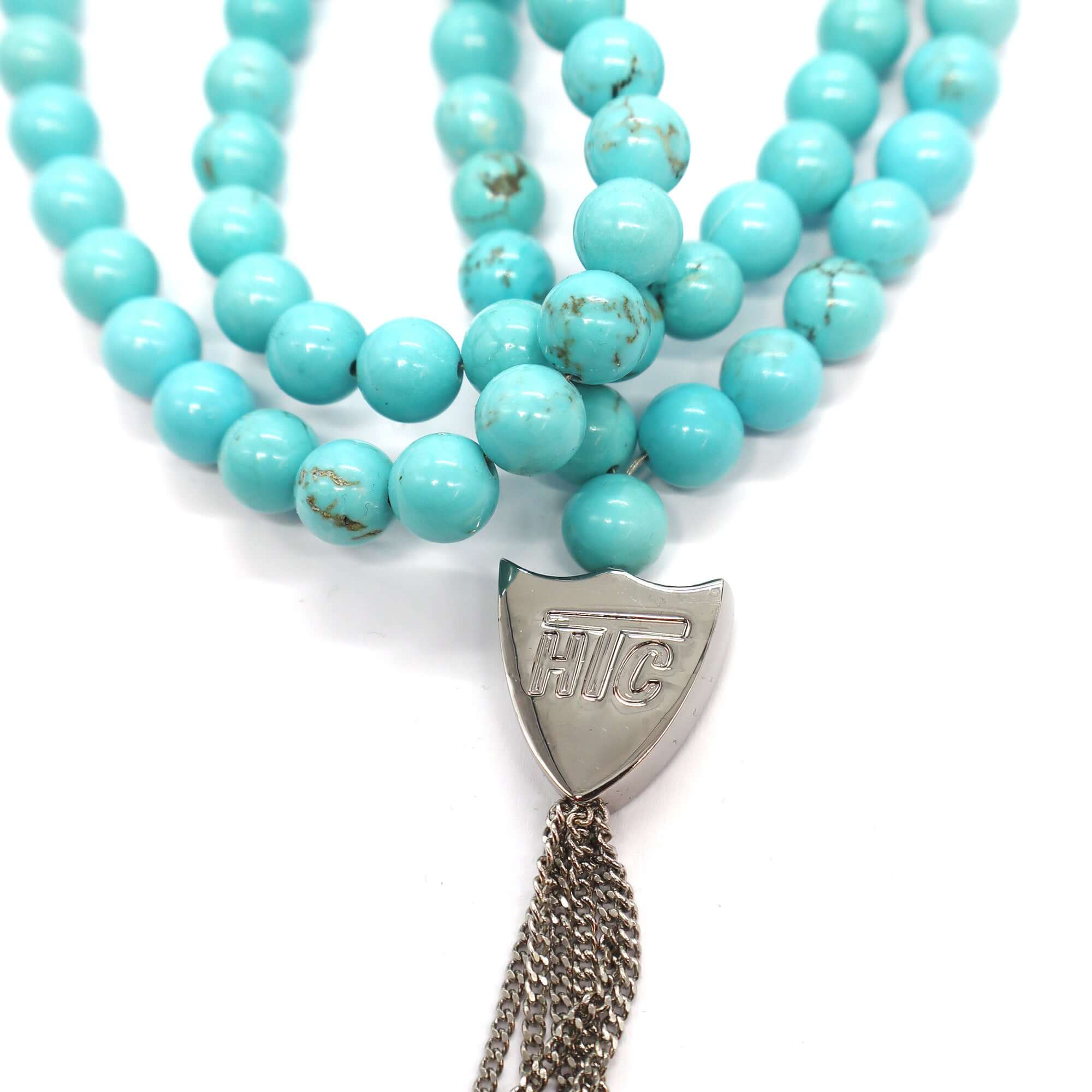 htc los angeles MYSTERY SHIELD NECKLACE TURQUOISE Turquoise necklace featuring a blend of textile, glass, and metal materials, with a metal HTC logo detail. Hollywood Trading Company
