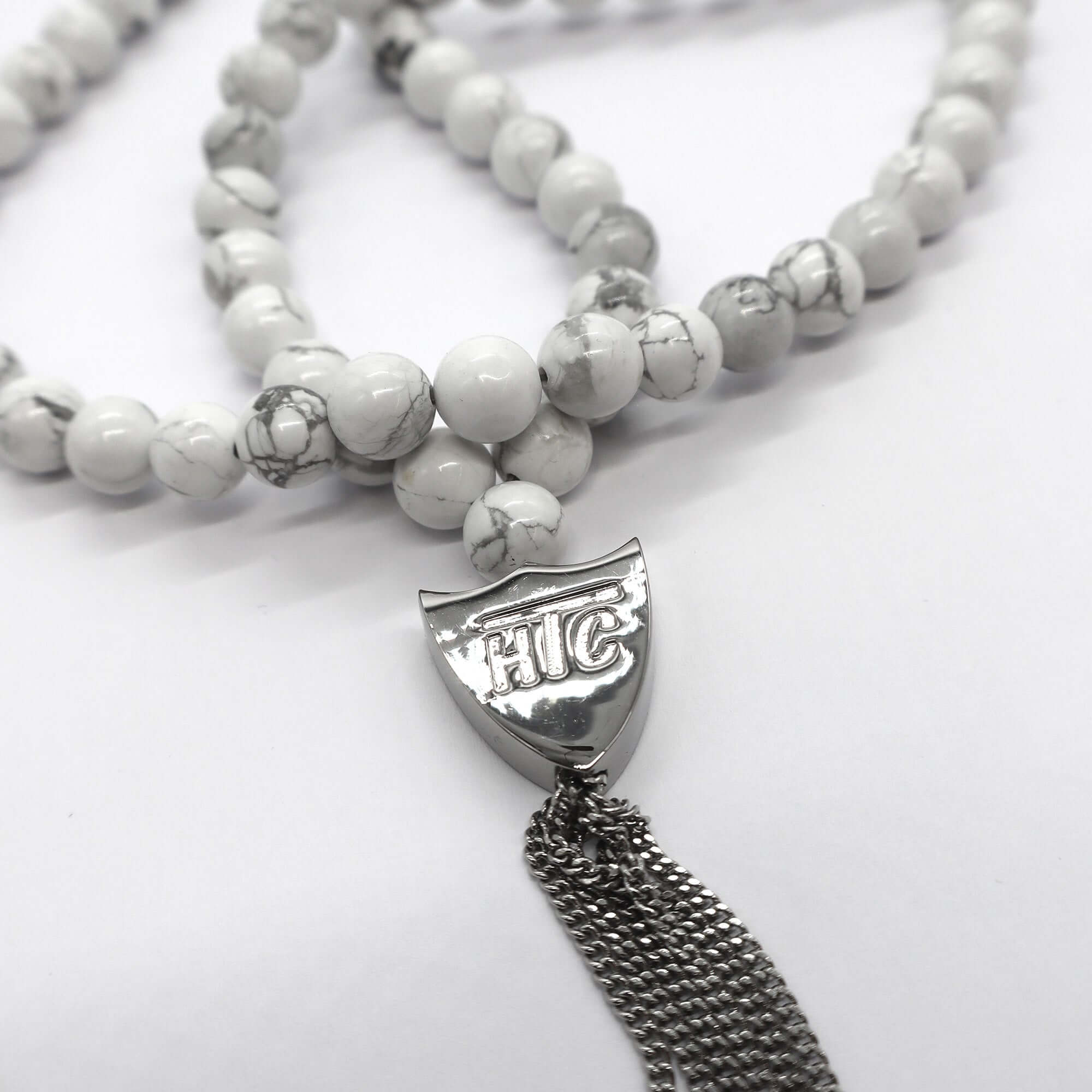 htc los angeles MYSTERY SHIELD NECKLACE WHITE White necklace featuring a blend of textile, glass, and metal materials, with a metal HTC logo detail. Hollywood Trading Company